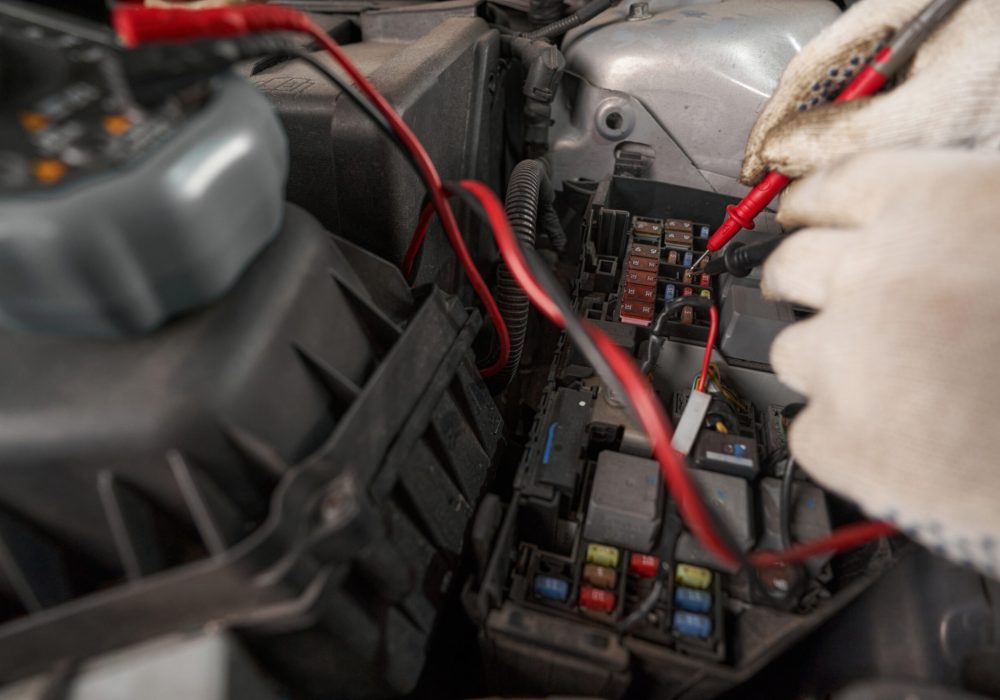 Man in blue protective clothes standing near car with open car hood and checking integrity of electric fuses
